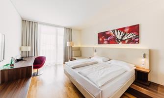 A modern hotel room with a large double bed and a desk. The room is bright and friendly, with a piece of artwork on the wall.