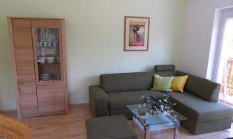 A cozy living room with a modern gray sofa and a glass table. A framed picture hangs on the wall and a bookshelf is visible.