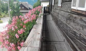 A wooden balcony with colorful flowers and a view of a green landscape. In the background, traditional houses and mountains can be seen.