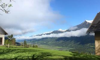 A picturesque view of the mountains with snow-capped peaks and dense clouds. In the foreground, there is a green meadow and rural buildings.