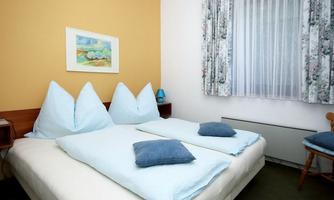 A cozy bedroom with a double bed and blue pillows. The walls are designed in soft color tones and a window with curtains lets in plenty of light.
