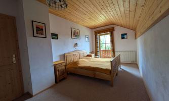A cozy bedroom with a wooden ceiling and decorative frames on the walls. A large window offers a view of the balcony and provides plenty of daylight.