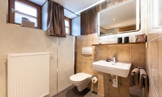 A modern bathroom with bright tiles and wooden details. It features a sink, a toilet, and windows for natural light.