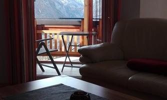 A cozy room with a sofa and a table. Through the window, one can see the mountains and a balcony.