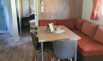 A modern living room with a dining table and comfortable chairs. The walls are made of wood and the room has large windows with orange curtains.