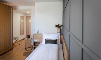 A modern bedroom with a single bed and a wooden wardrobe. The room has bright walls and a minimally designed table.