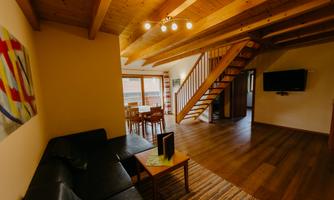 A modern living room with a dark couch and a dining area. A wooden staircase leads to the upper floor, and large windows provide plenty of daylight.