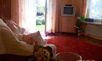A cozy living room with a beige sofa and a red carpet. Light streams in through the windows, and in the background, a TV is visible.
