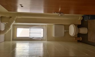 A small bathroom with a toilet and a window. The room has light walls and a wooden cabinet.