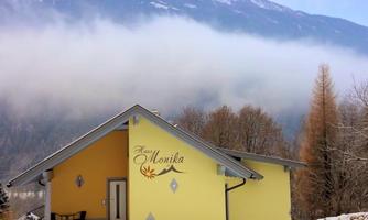 A yellow holiday home named "Haus Monika" stands in a wintry landscape. In the background, snow-covered mountains and fog can be seen.