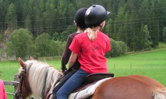 Two children are riding on a horse in a green landscape. In the background, there are trees and a beautiful hill.
