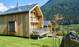 A modern wooden house in the mountains with a green meadow in front. The surroundings are picturesque with trees and mountains in the background.