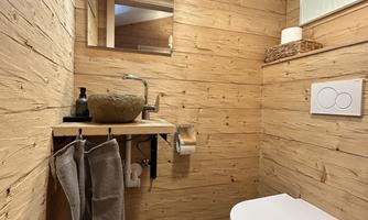 A modern bathroom with wooden walls and a large mirror. It features a stylish toilet and a practical shelf for towels and toiletries.