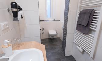 A modern bathroom with a sink, a toilet, and a towel holder. The walls are tiled in a light color and the floor is dark.