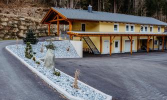 A modern building with a yellow facade and wooden cladding. The parking lot is paved, and there is a small greenery with stones and trees.