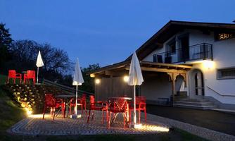 A cozy terrace with red furniture and umbrellas, illuminated by the evening light. In the background, a modern house can be seen.