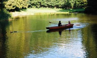 A man and a woman are rowing in a boat on a calm lake. Surrounded by trees, the landscape is reflected in the water.