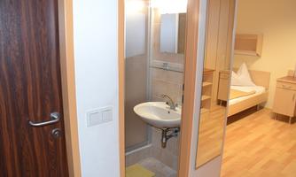 A simple room with a sink and a mirror. In the background, a bed and a wooden floor are visible.