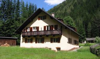 A beautiful multi-story house surrounded by green meadows and trees. In the background, mountains are visible and the sky is blue with some clouds.