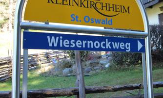 A signpost in Bad Kleinkirchheim indicates the direction to Wiesernockweg. In the background, typical mountain landscapes can be seen.