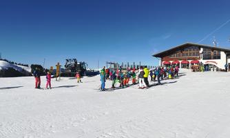 A group of skiers is standing in line at a ski lift. In the background, delicious sun loungers and a blue sky can be seen.