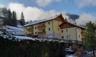 A picturesque residential building in the mountains, surrounded by light snow. The clear blue sky and the trees create a cozy atmosphere.