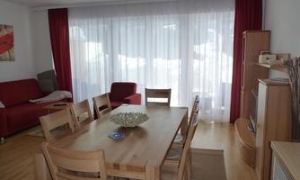 A bright living room with a large wooden dining table and chairs. In the background, there is a red sofa and large windows with curtains.