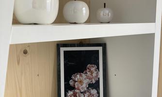 A modern shelf corner with white ceramic apples and a framed picture of flowers. The background is made of light wood, creating a cozy atmosphere.