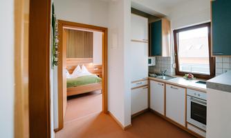 A modern kitchen with white cabinets and wooden shelves. In the background, there is a bedroom with a double bed.
