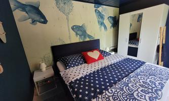A modernly furnished bedroom with a large bed and a striking wallpaper featuring fish. There is a white wardrobe and bedside table that complement the room.