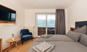A modern room with two beds, a cozy armchair, and a television. Large windows offer a beautiful view of the mountains.