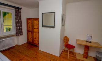A simple room with wooden floors and neutral colors. There is a table, a chair, a wardrobe, and a window with curtains.