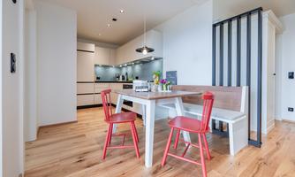 A modern dining room with a table and red chairs. The kitchen in the background is bright and well-equipped.