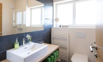 A modern bathroom with a white sink and large windows. There is a bright and friendly atmosphere with green towels and plants.