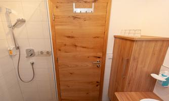 A modern bathroom with a shower and a wooden door. The sink area features a wooden shelf and a clear, functional style.