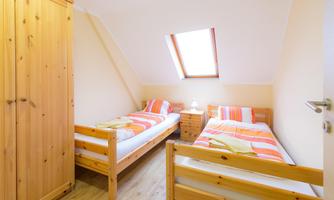 A simple bedroom with two single beds and a wooden wardrobe. The room has a window with daylight and orange-white bed linen.