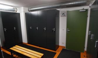 A changing room with black lockers and a wooden bench. The walls are light, and the door is green.