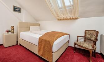 A cozy bedroom with a single bed and a chair. The room has a red carpet and large windows with curtains.