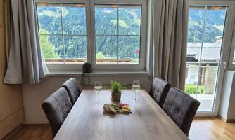 An inviting dining area with a large wooden table and comfortable chairs. Through the windows, one looks out at a picturesque mountain landscape.