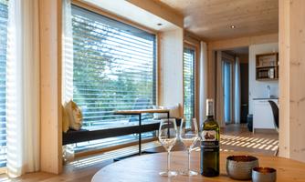 A modern living room with large windows, transparent light, and a dining table. On the table are two wine glasses and a bottle of wine, surrounded by cozy furnishings.