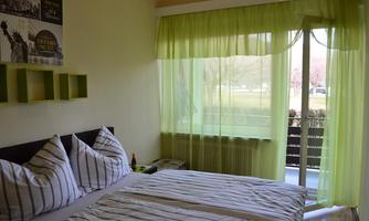 A bright bedroom with white bedding and striped pillows. Large windows with green curtains let in plenty of daylight.
