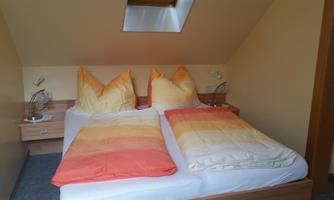 A cozy bedroom with a double bed and large, colorful cushions. The walls are light yellow and there are modern bedside lamps.
