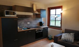 A modern kitchen with black cabinets and a stove. Next to it is a cozy sofa and a table in a bright room.