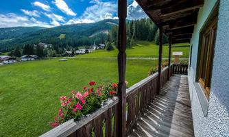 A balcony with a view of a green meadow and wooded mountains. Colorful flowers decorate the railing of the balcony.