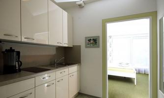 A modern kitchen with light cabinets and a cooktop. In the background, a bedroom with a bed and plenty of light is visible.