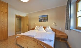 A cozy bedroom with a double bed and wooden furniture. A picture hangs on the wall, and there are large windows that let in a lot of light.