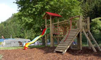 A playground with a wooden structure, slide, and climbing elements. The area is surrounded by trees and has a soft ground made of bark mulch.
