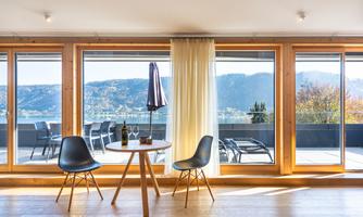 A bright room with large windows and a view of the mountains. In the foreground, there is a round table with two chairs and a bottle of wine.