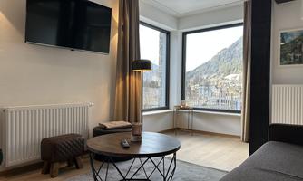A modern living room with a TV on the wall and a round table. Large windows offer a view of the mountains.