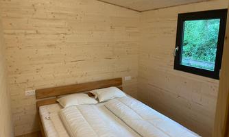 A simple bedroom with a double bed and light wooden walls. A window lets in natural light and offers a view of greenery.
